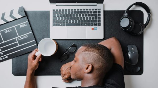 Top down photo of a work desk with a laptop, headphones, mouse, glasses, clapper board, with a person sleeping holding a mug.
