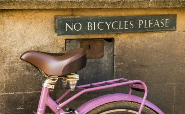Photo of a sign on a building that says “No Bicycles Please” with a pink bicycle right under the sign.