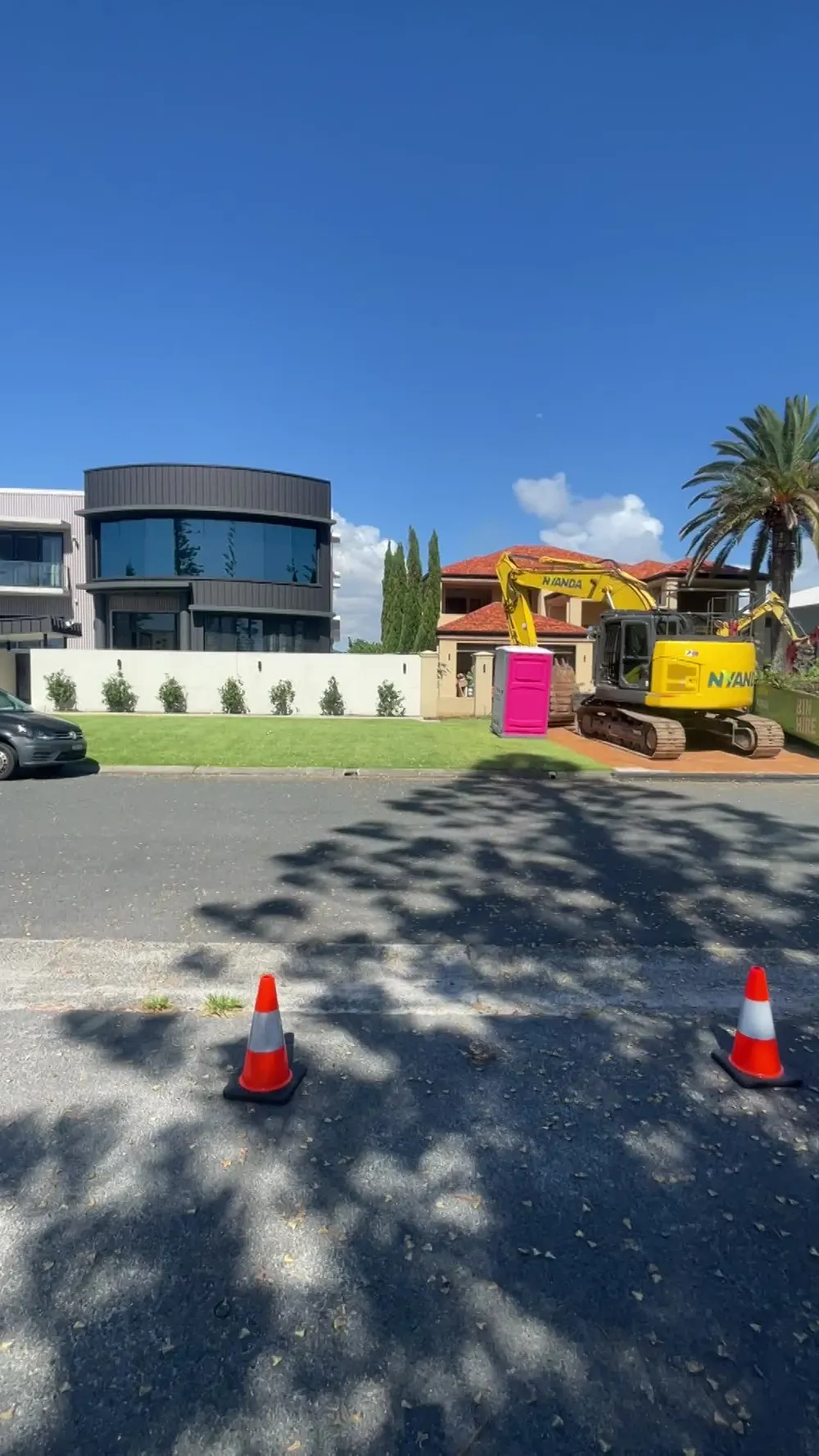 Yellow excavator and pink portable toilet on a canal-front lot on demolition day one, traffic cones on the street