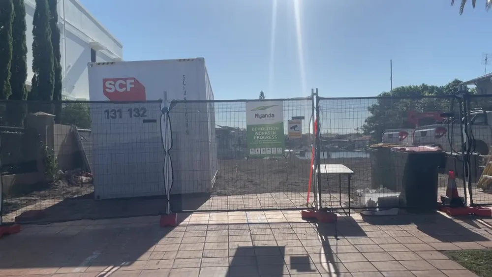 Fenced vacant building site with a site office container, demolition sign and wheelie bins on the footpath