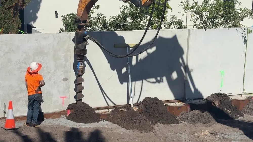 An auger on an excavator pre-drilling a dewatering spear hole beside the steel sheet piling and the new precast boundary fence