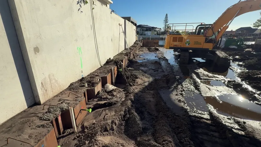 Dewatering spear risers in the sheet pile pans along the new precast boundary fence, with a muddy, waterlogged site and an excavator beyond