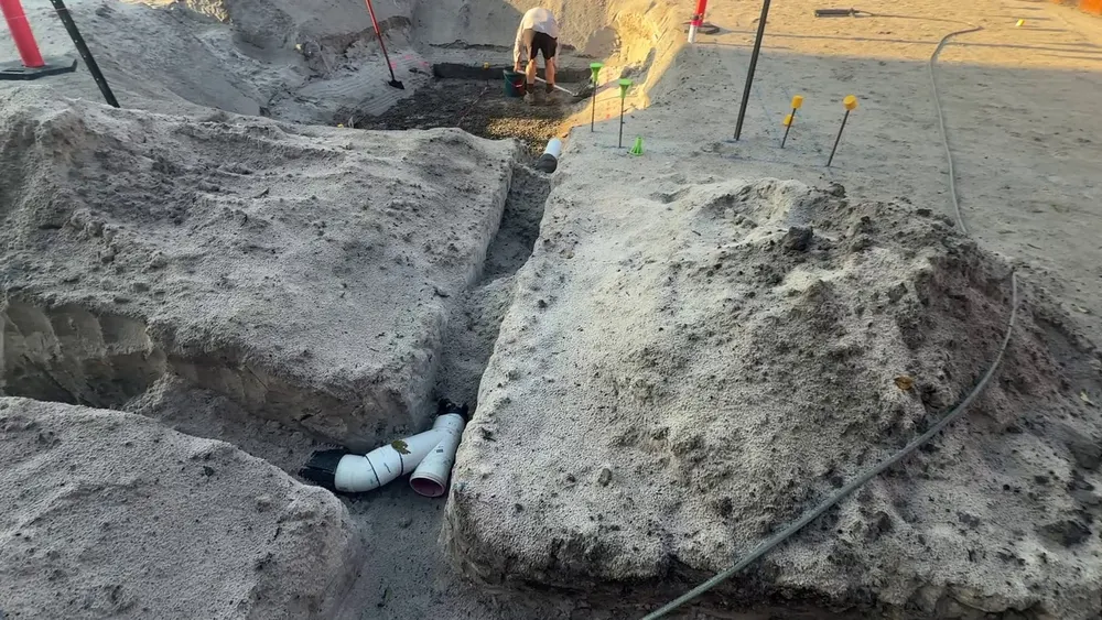 A hand-dug trench in white sand with the first sheathed sewer pipe fittings, and capped height pins set out for the blinding behind