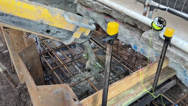 Concrete pouring from a truck chute into the reinforcing steel cage of a boundary fence footing, between steel sheet piling and a string line