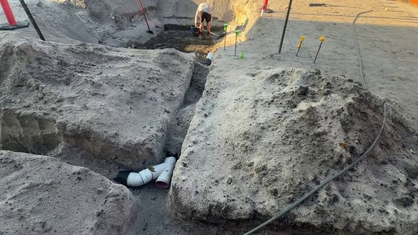 A hand-dug trench in white sand with the first sheathed sewer pipe fittings, and capped height pins set out for the blinding behind