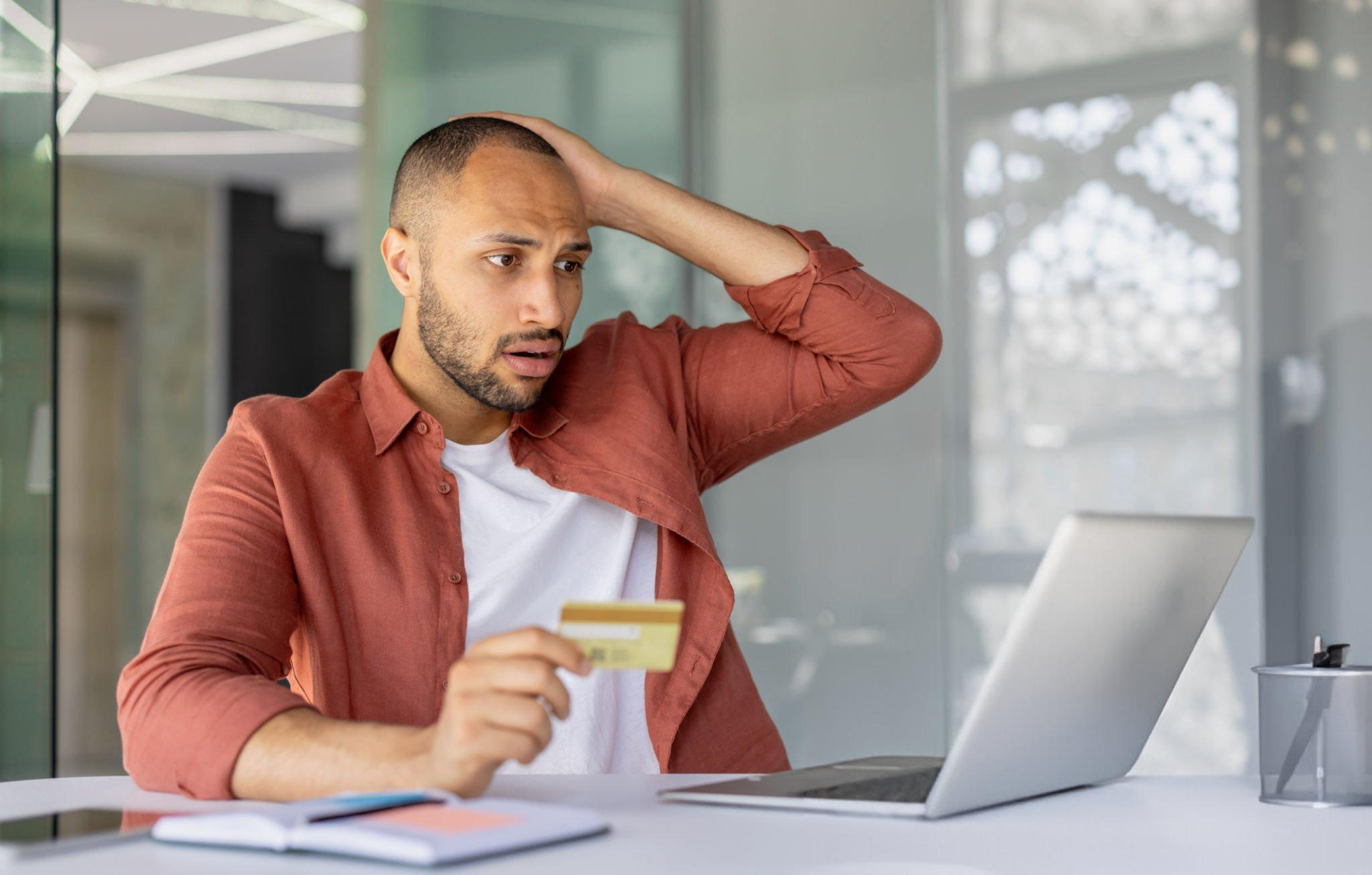 A man holding a credit card and in shock as he can see from his PC that his credit has been frozen