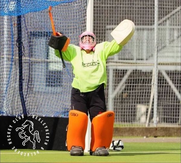 Hockey goalkeeper in full kit, arms aloft smiling at the camera. The smock is bright green. 