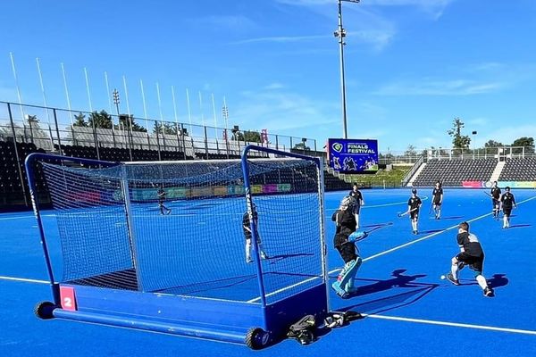 Team Bexley in black tops warming up on the blue hockey pitch. The pitch is surrounded by stadium seating. It's also a sunny day with almost clear blue skies