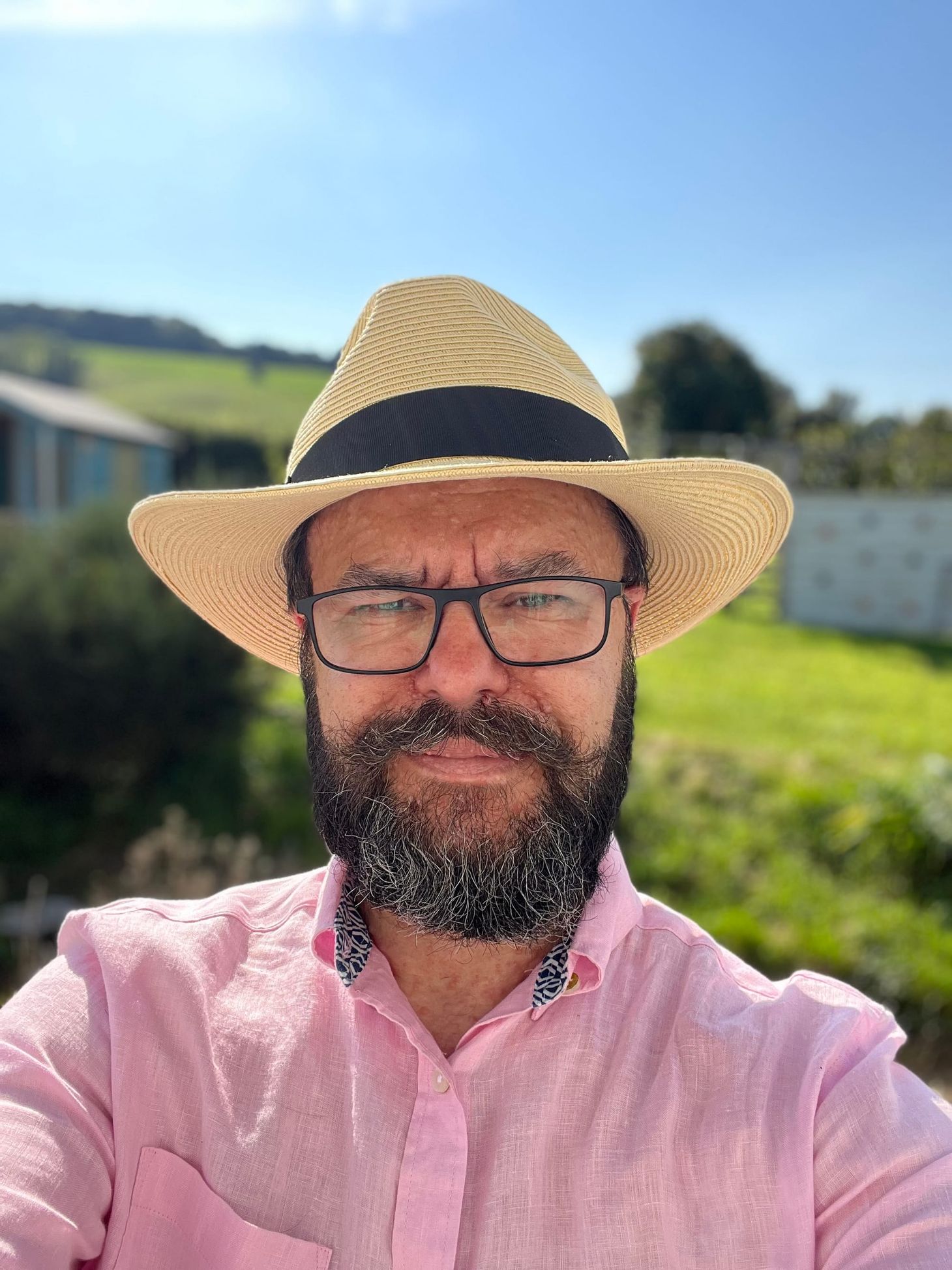 A bearded man with glasses grins coquettishly at the camera. He is wearing a pink shirt and a straw fedora. 