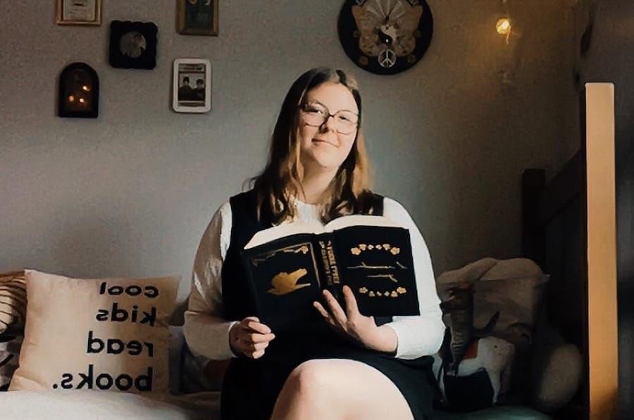 Courtney sits on a bed with a book open in hand, looking and smiling at the camera.