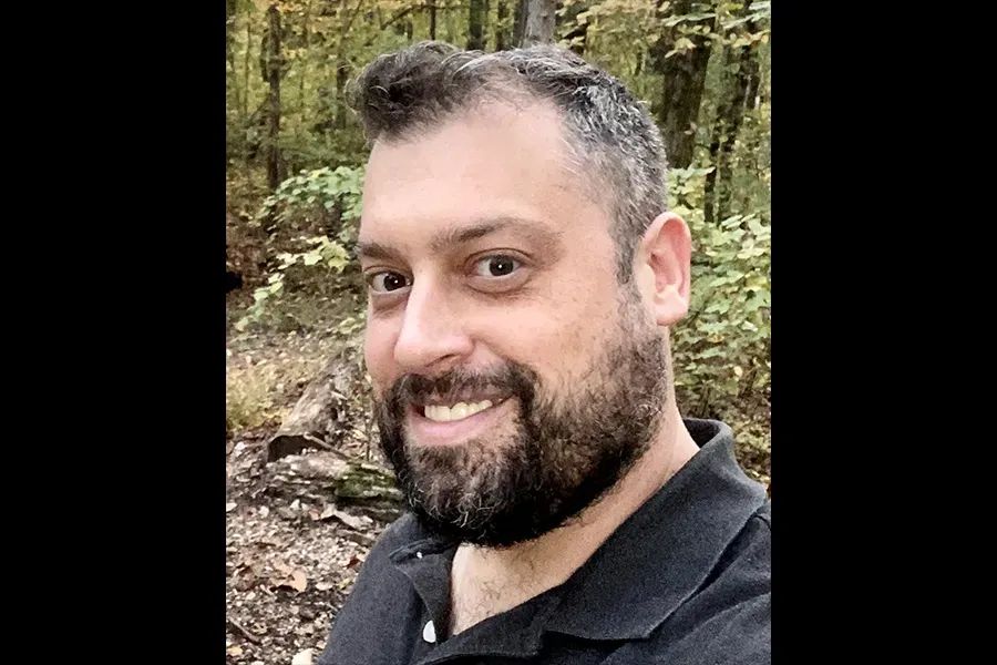 Poet Joshua St. Claire is standing in a forest. He is smiling with a brown beard.