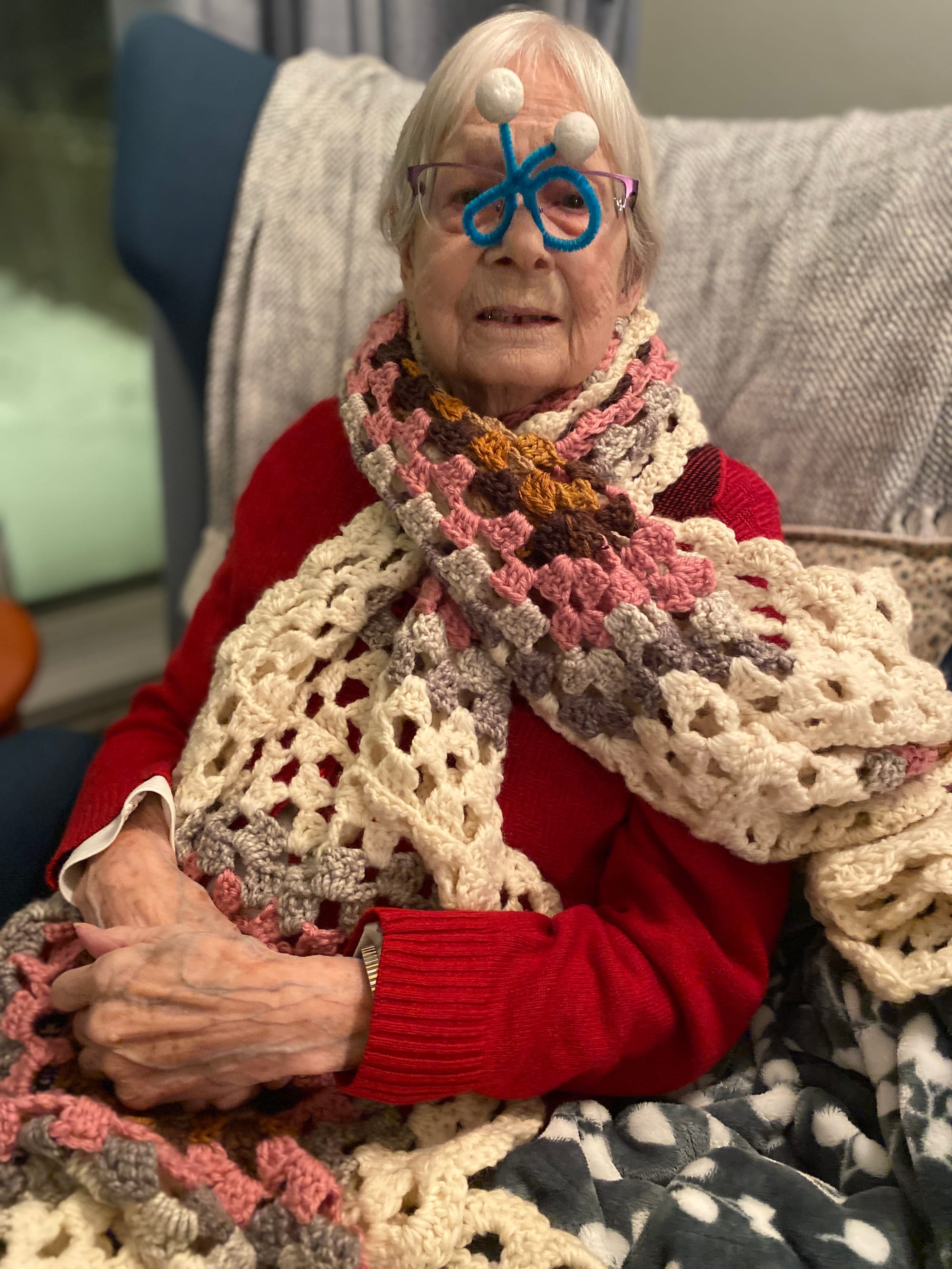 A 91 year old woman with short white hair wears a crocheted shawl with a blue pipe cleaner shaped into glasses with styrofoam balls like antenna on top, balanced on her real glasses
