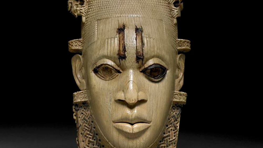 A mask in yellow white ivory with a broad nose and lips one blacked out eye, two dark rectangles between the eyes against a back background