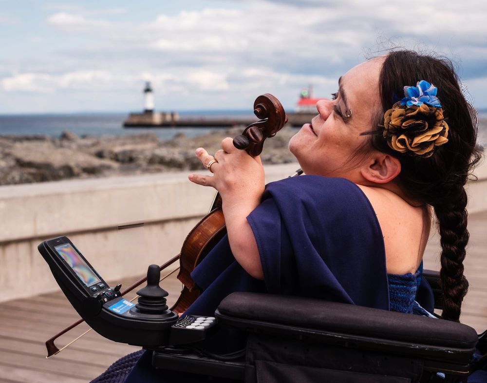 A woman with a long braid in a purple dress plays a violin like a cello sitting a wheel chair smiling into the wind, in the distance are the rocks and a lighthouse and the great Lake Superior