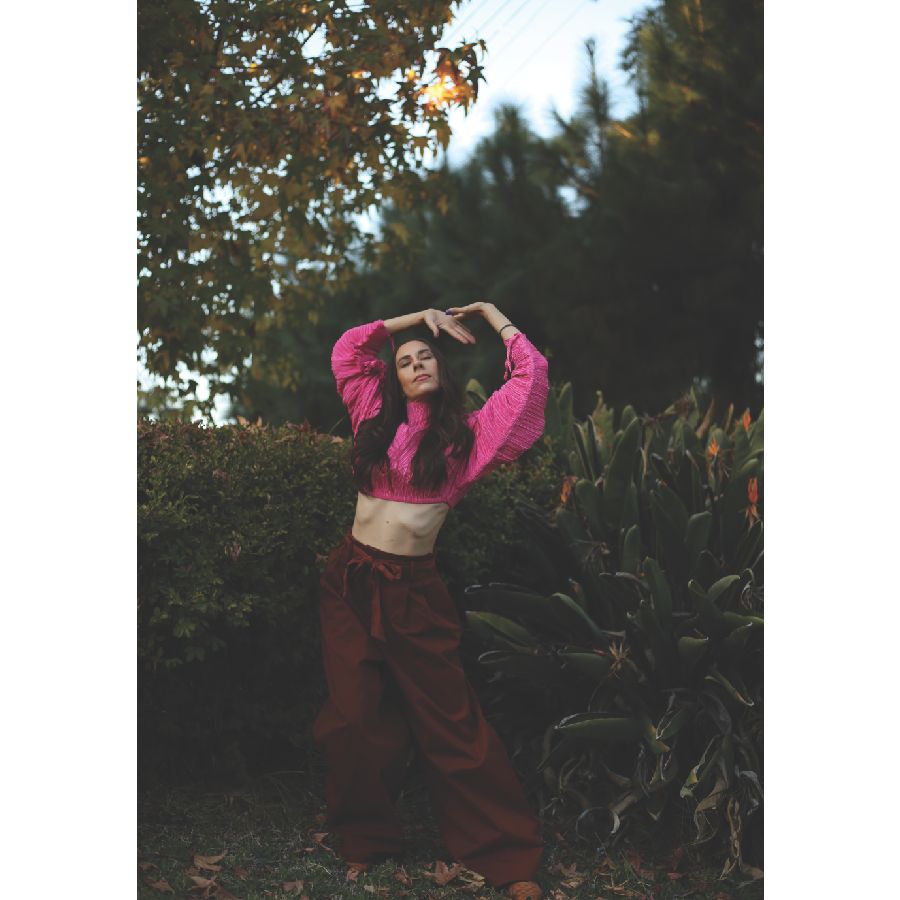 a white woman with long brown hair stretches her arms over her head in a bright pink long sleeved crop top, eyes closed again
