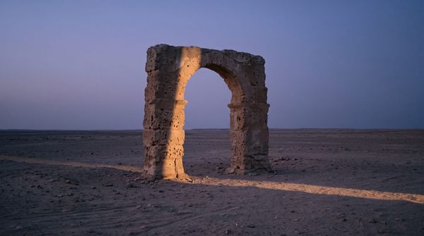 Solitary ancient limestone archway standing in a vast, minimalist desert landscape at dawn with dramatic directional light.