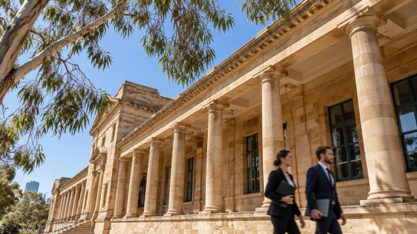 Low-angle view of a sandstone heritage building colonnade with eucalyptus branches and a clear blue sky, two people walking past.