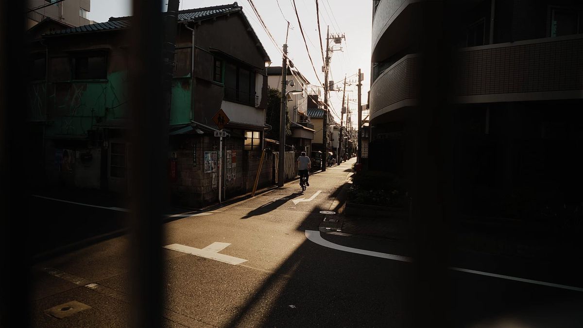 Un hombre en bicicleta por una callejuela de Tokio, con postes y cables, bajo una luz rasante que deja la calle entre sol y sombra