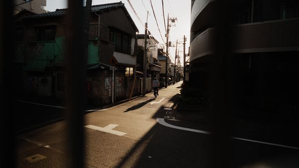 Un hombre en bicicleta por una callejuela de Tokio, con postes y cables, bajo una luz rasante que deja la calle entre sol y sombra