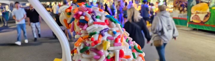 vanilla soft serve ice cream cone with rainbow sprinkles at a county fairground at night