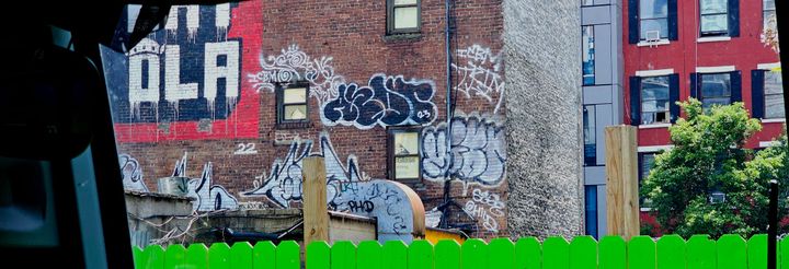 View from a bus: a green fence, a brick tenement building wall covered in graffiti, a tree. 