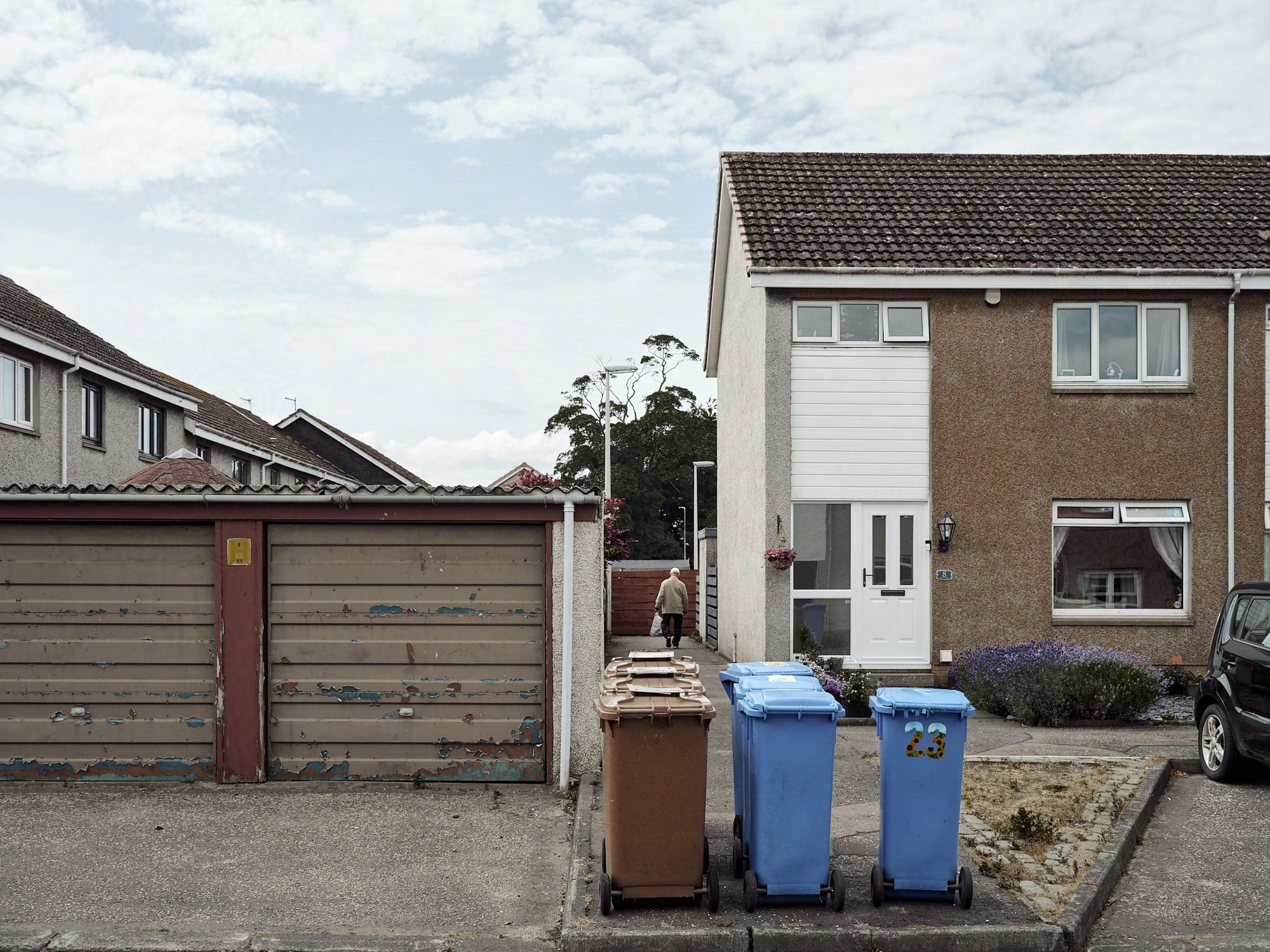 A colour image of a neat grid of wheelie bins. To one side is an old garage lock-up, paint flecking of the doors, behind that the roofs of surburban house trail of in to the background. To the other side another house stand large over proceedings, while in the middle, an old man carries his shopping behind the bins and down a narrow alleyway.