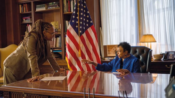 Two people face each other across a polished office desk, one leaning forward and the other seated gesturing, with a U.S. flag behind them.