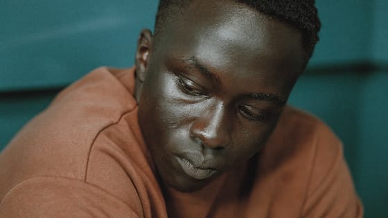 Close-up of a young Black man in a rust-brown sweatshirt looking down pensively against a blurred teal background.