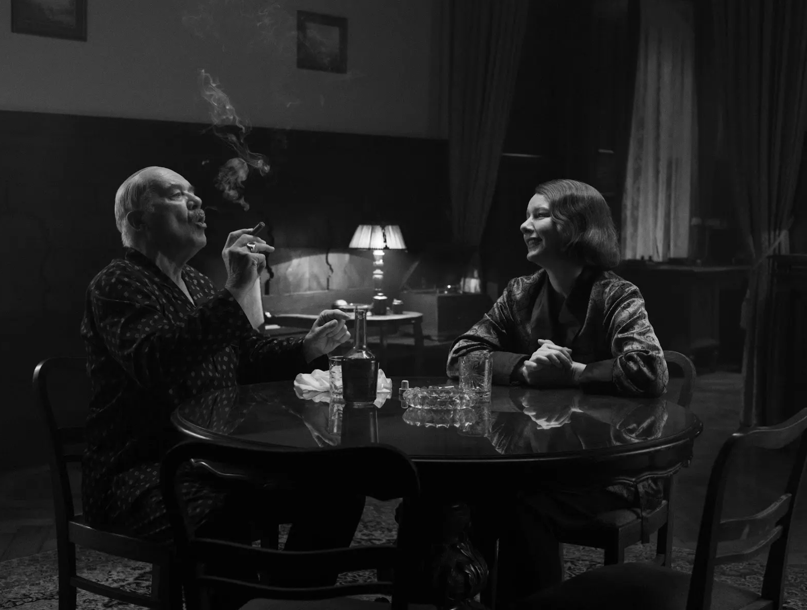 Black-and-white still of an older man smoking a cigar and a smiling woman seated at a round table in a dim, vintage room.