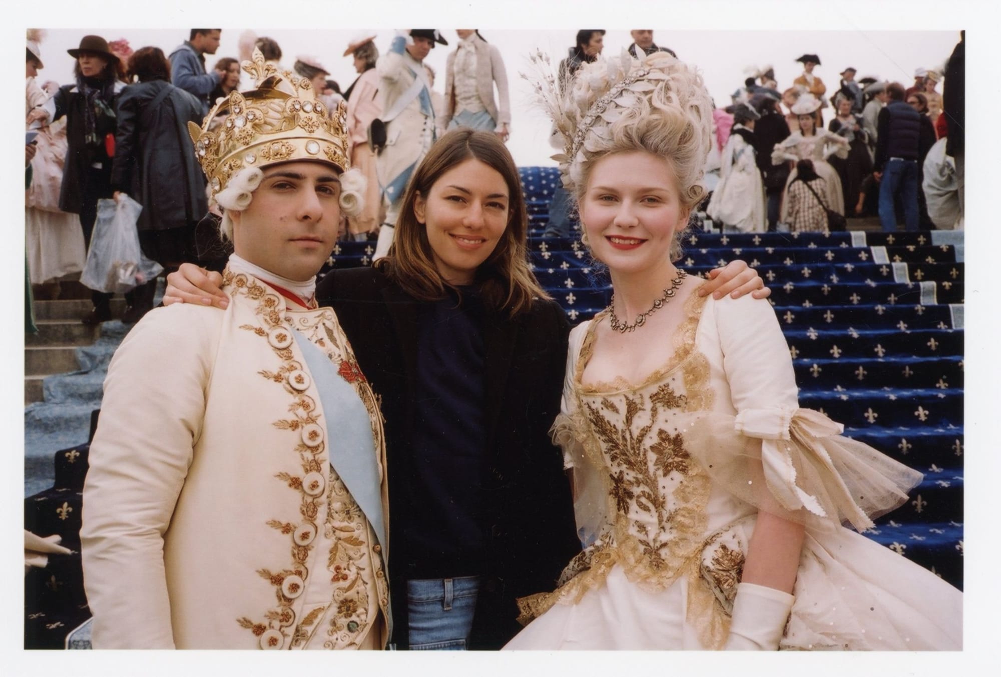 Sofia Coppola stands between two Marie Antoinette actors in ornate eighteenth-century costumes on blue fleur-de-lis steps.