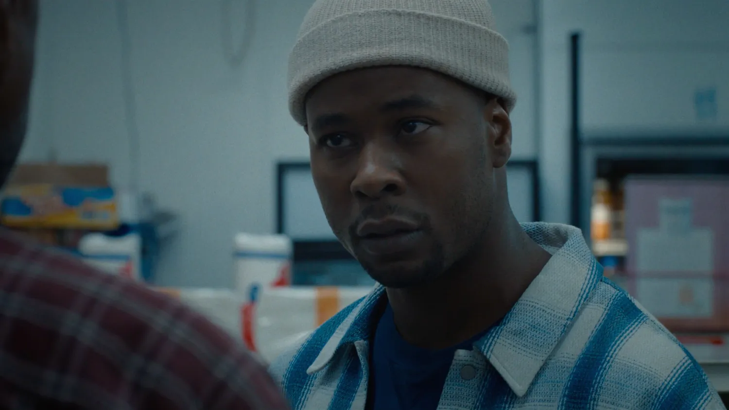 Film still from Spilled Milk showing a man in a cream beanie and blue plaid overshirt facing someone in a muted indoor setting.