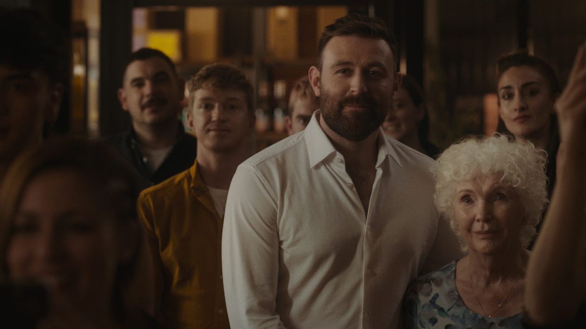 Group of people in a dimly lit bar, centered on a bearded man in a white shirt, flanked by a younger man and elderly woman.