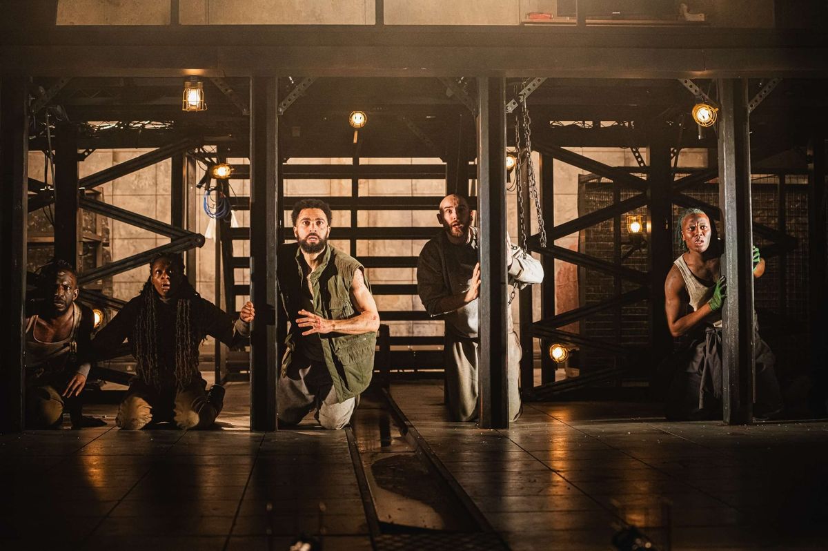Five actors crouch tensely on a dark industrial stage set with metal beams, chains, and warm lantern lighting during a theater production.