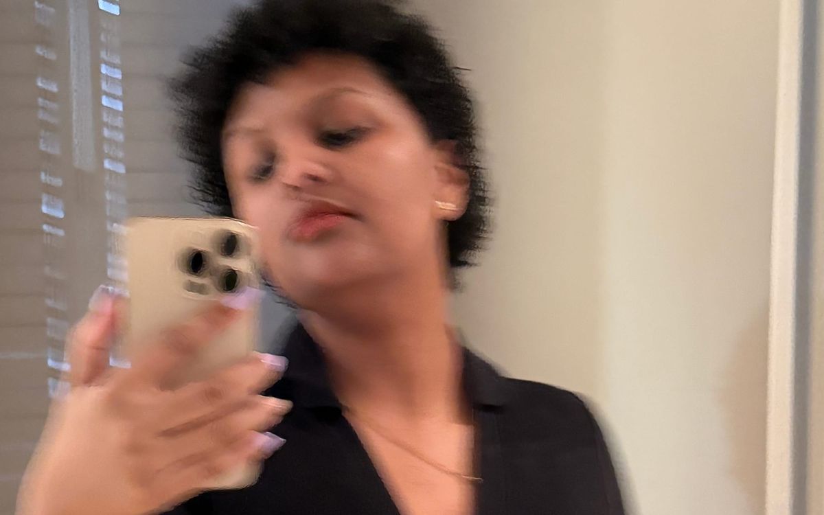 Blurry mirror selfie of a woman with short curly black hair, red lipstick, black top, holding a gold iPhone with long nails.