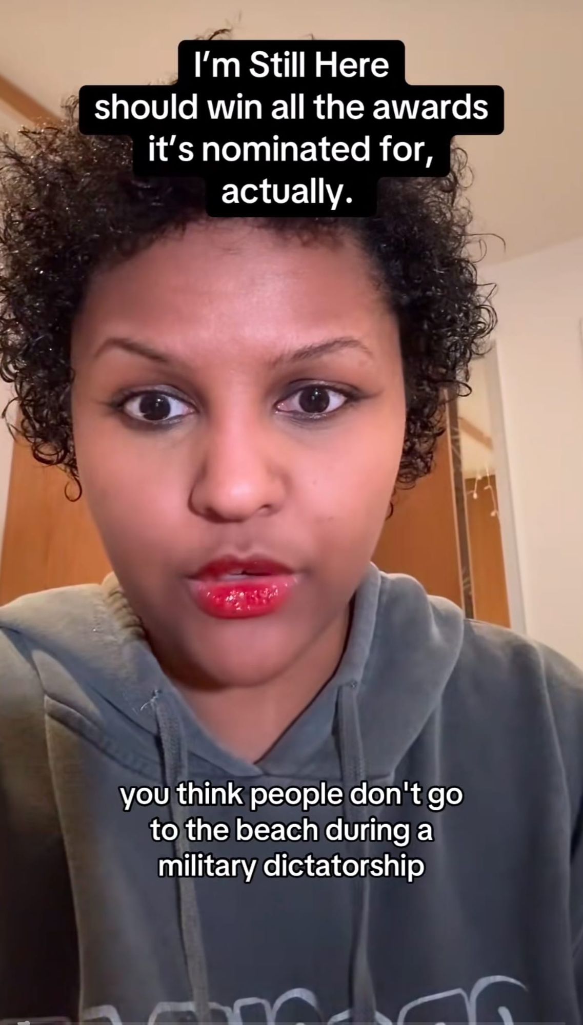 Woman with curly hair and red lipstick in gray hoodie, with text overlay praising film "I'm Still Here" and discussing beach during military dictatorship.