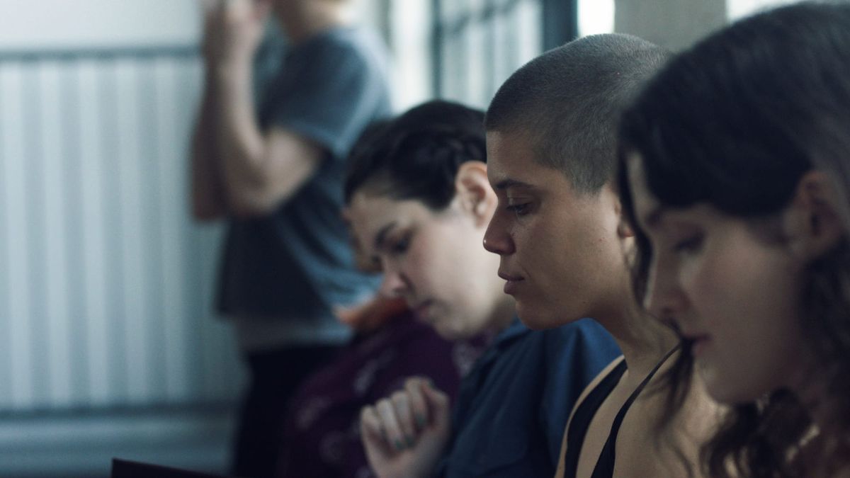 Several young people sitting in a row with downcast eyes; a person with a buzzcut in focus in the center, dimly lit room.