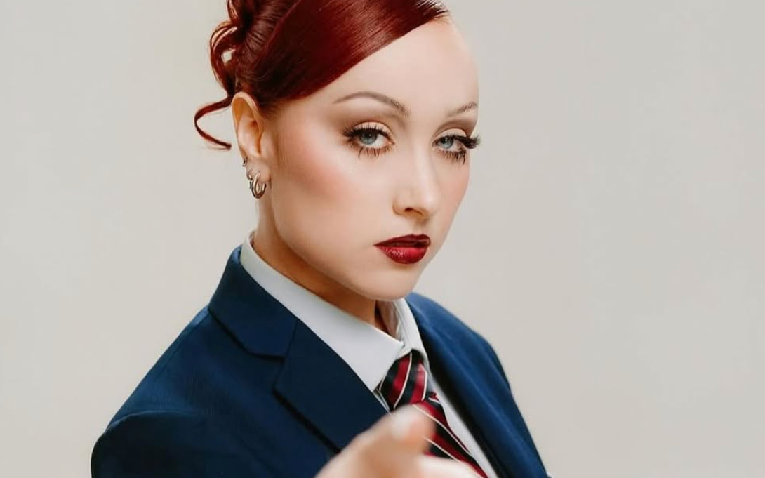Young woman with red updo hair, blue eyes, red lipstick, wearing navy suit with striped tie, pointing toward camera.