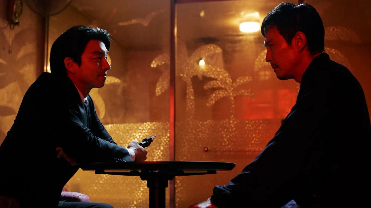 Two men in dark clothing face each other across a small table in a dimly lit bar with warm amber and red lighting.