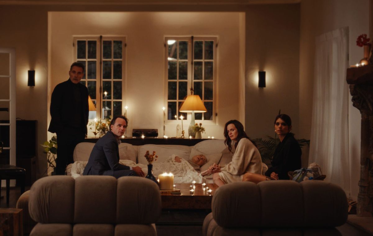 Five people in a candlelit living room; four sit or stand around an elderly person lying on a couch, appearing somber.