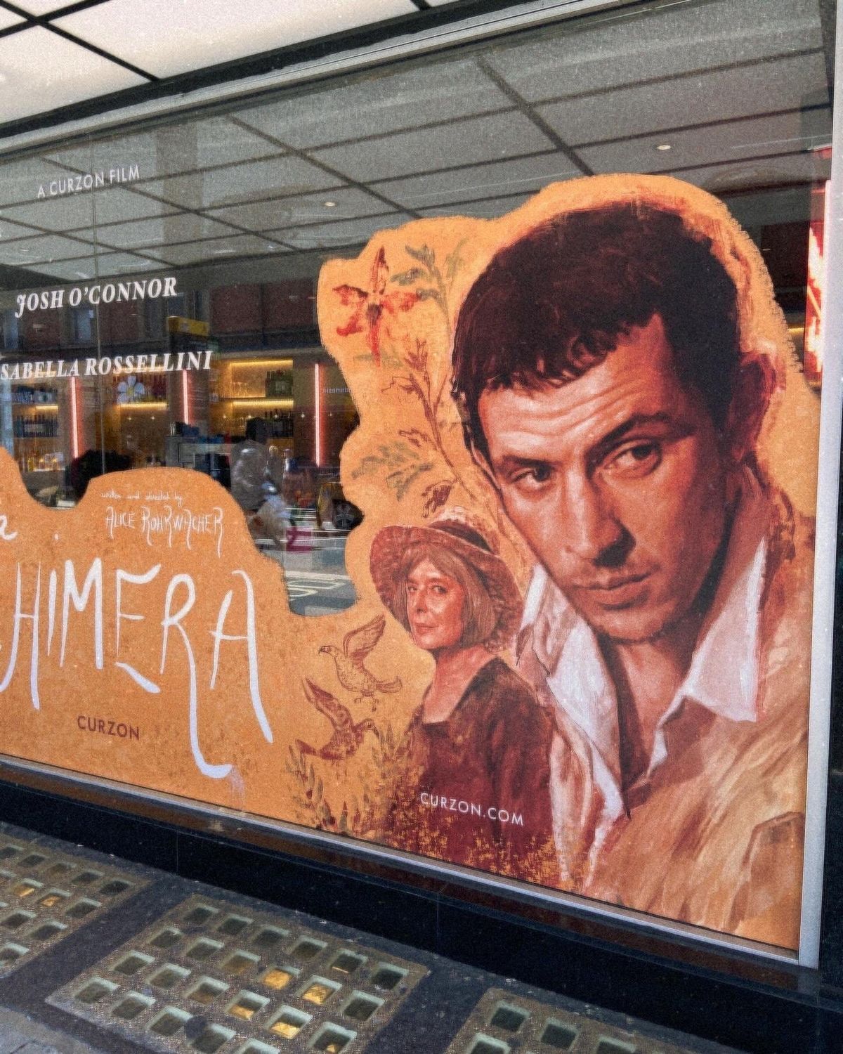 Movie poster for "La Chimera" in a window, featuring painted portraits of Josh O'Connor and Isabella Rossellini, by Alice Rohrwacher.
