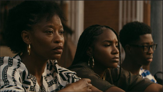 Three Black people sitting together looking somber; a woman in a patterned blouse, a young woman with braids, and a young man in glasses.