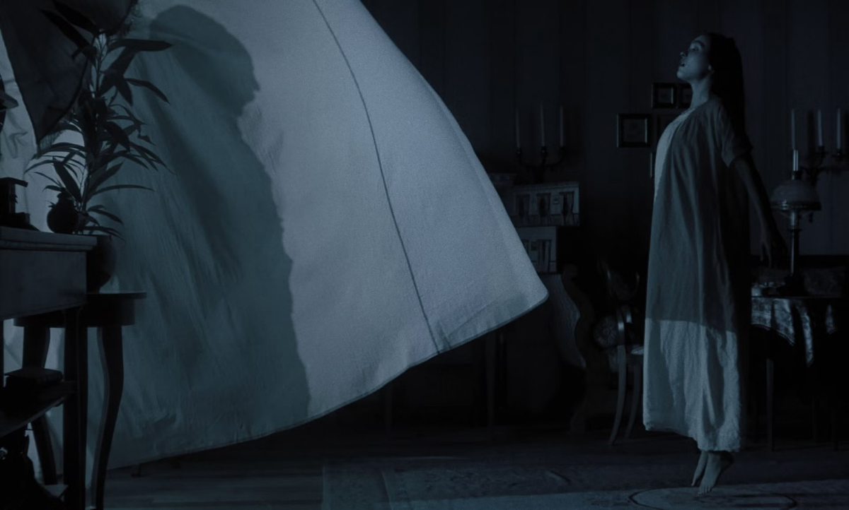 Dark room with woman in white nightgown standing barefoot, looking up at a large white sheet billowing eerily in the air.