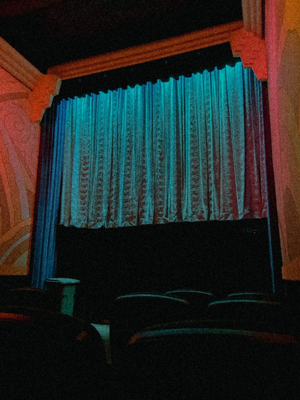 Dimly lit vintage movie theater with teal curtain drawn over the screen, ornate Art Deco walls, and empty seats below.