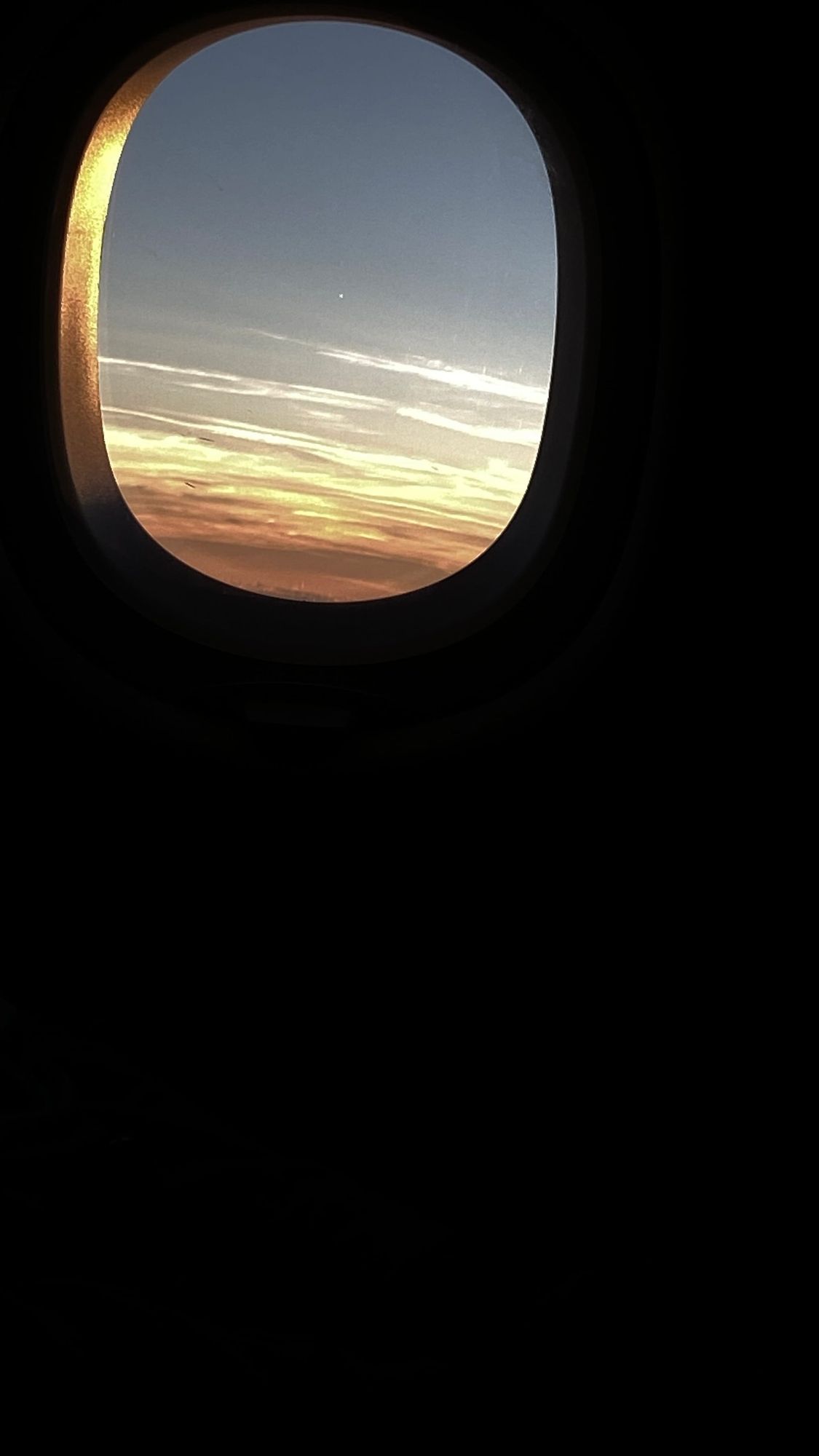 View through an airplane window showing a sunset with orange, yellow, and pink clouds against a pale blue sky, cabin dark.