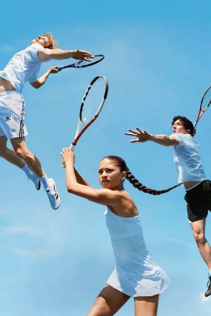 Three tennis players in white athletic wear swing rackets against a blue sky, shot from a low angle looking upward.