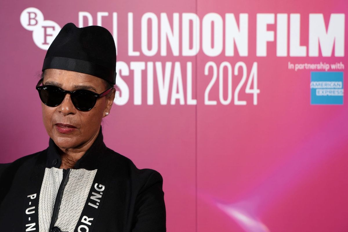 Person in black hat, sunglasses, and black blazer posing at BFI London Film Festival 2024 pink backdrop.
