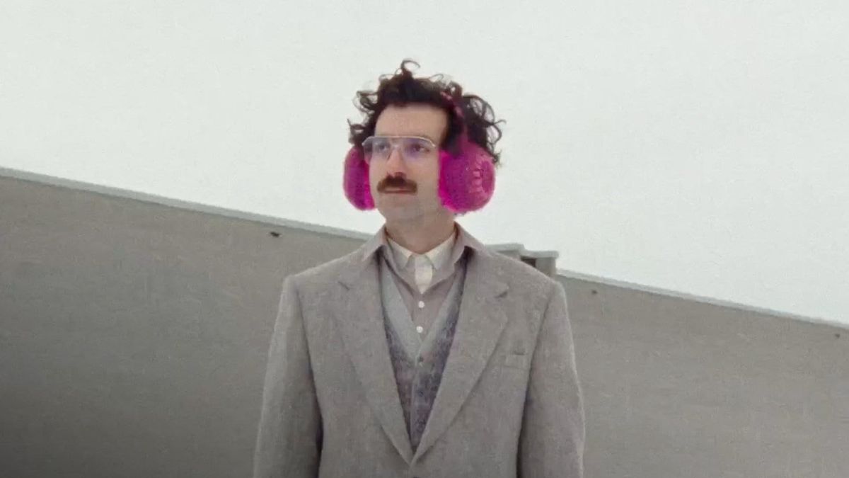 Man with curly dark hair, mustache, glasses, and pink earmuffs wearing a gray suit, standing against a white wall.