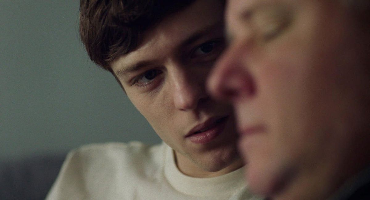 Close-up of a young man with dark brown hair and a white t-shirt looking intently at another person whose face is partially visible in the foreground.