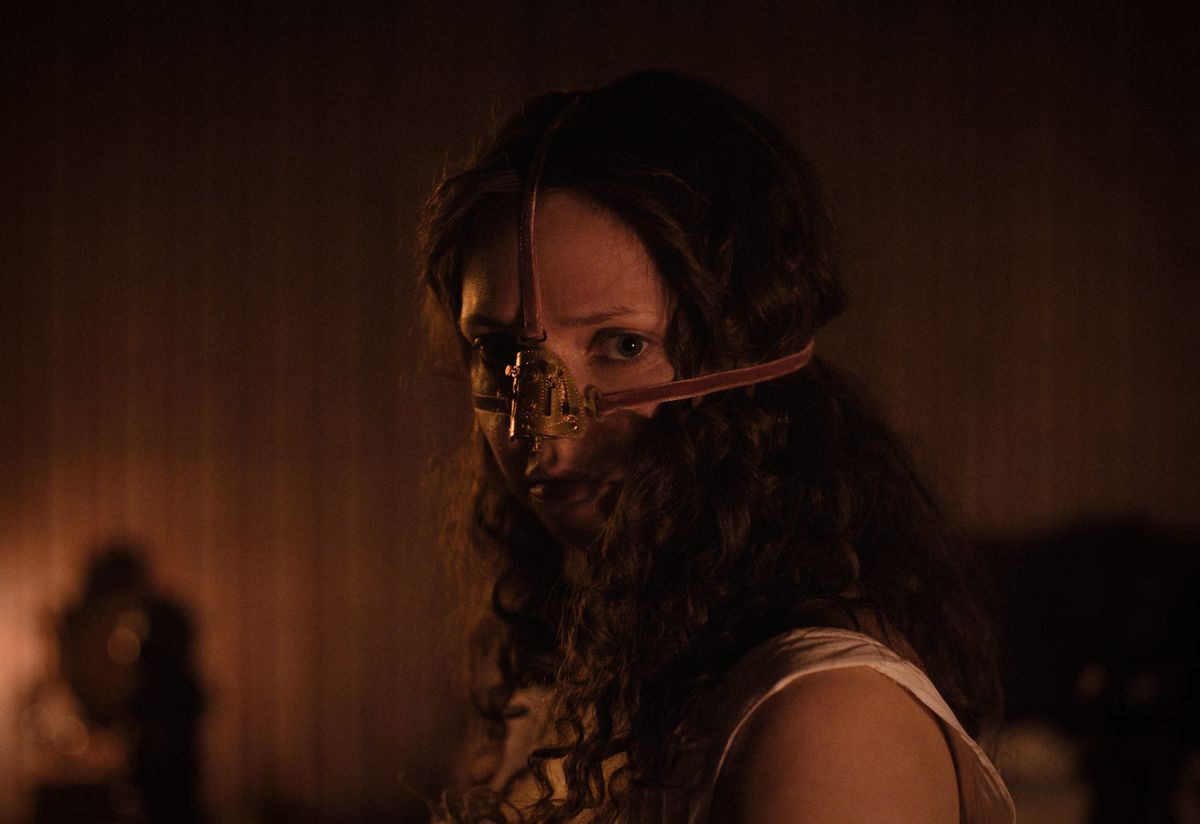 Woman with long curly hair wearing a metal nose prosthetic or mask device with leather straps, lit in warm amber tones.