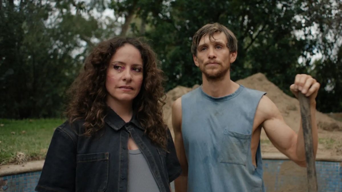 Two people standing outdoors: a woman with curly dark hair in a denim jacket and a man in a blue sleeveless shirt holding a shovel.